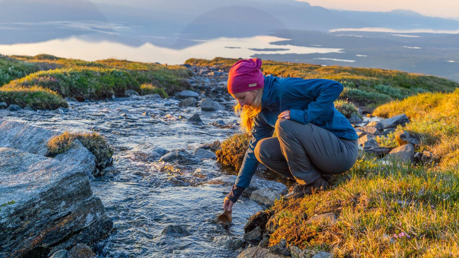 Kvinna dricker vatten ur bäck med Torneträsk i bakgrunden. Abisko nationalpark, Sverige.