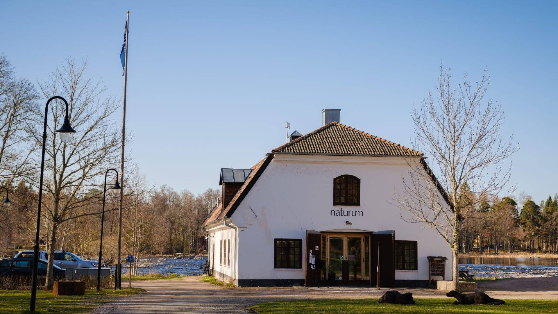Byggnaden som rymmer naturum ligger vid en grusad gångväg omgiven av träd och öppet landskap under en klar blå himmel.
