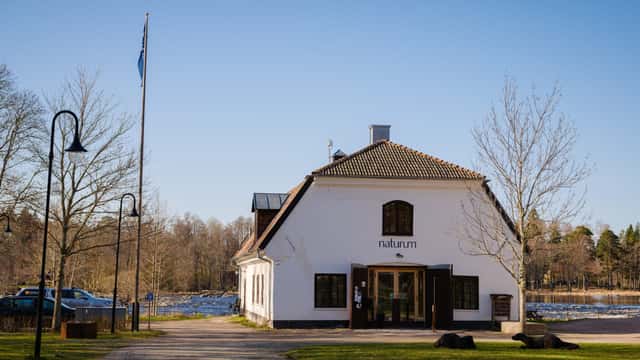 Byggnaden som rymmer naturum ligger vid en grusad gångväg omgiven av träd och öppet landskap under en klar blå himmel.